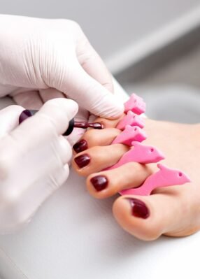 manicure-master-is-painting-female-toenails-with-maroon-nail-polish-by-brush-wearing-white-gloves manicure-master-is-painting-female-toenails-with-maroon-nail-polish-by-brush-wearing-white-gloves